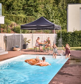 The black folding gazebo stands by the pool in the garden. Friends sit under it in the shade and have a pool and garden party.