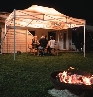 The 4.5x3 m garden tent stands in the garden in the dark. The 4 guests sit under it. There is a fireplace in front of the gazebo.