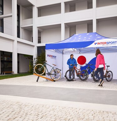 The printed promotion tent from "Intersport" stands on the outdoor area of the fair. Under the 4.5x3 m folding gazebo stands the salesman and his bicycles.